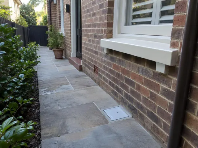 Dry paved side path beside a brick house with a drain grate in place in Double Bay